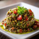 A close-up of a molded portion of vibrant lentil salad mixed with diced tomatoes and herbs, drizzled with olive oil.
