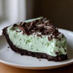 A close-up of a slice of creamy, light green grasshopper pie with a dark chocolate crust, topped with chocolate shavings.
