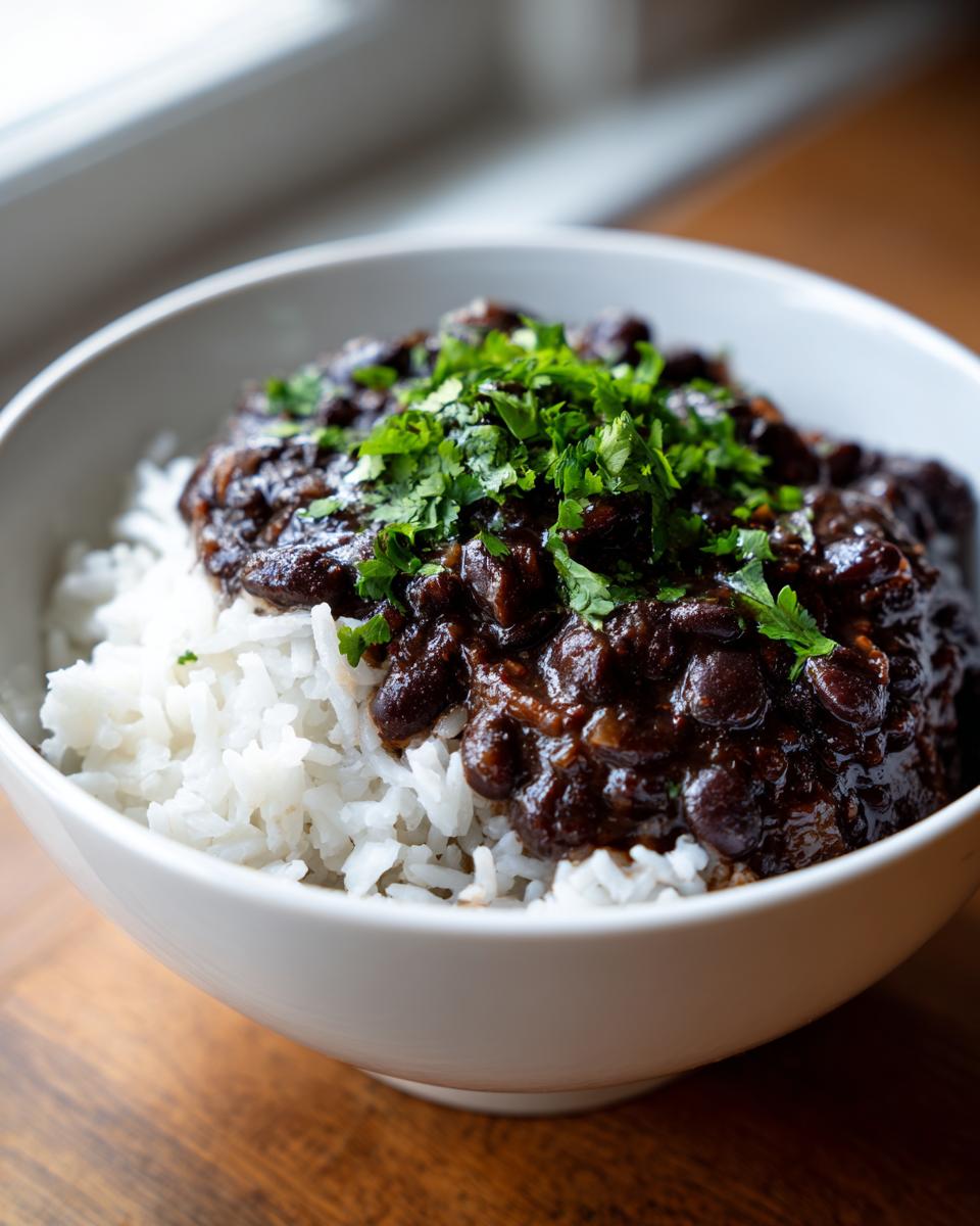 A close-up of a white bowl filled with white rice topped generously with rich, dark black beans and rice, garnished with fresh cilantro.