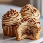 Three pecan pie cupcakes, one cut open to show the gooey filling and topped with brown sugar frosting and chopped pecans.