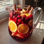 A clear glass pitcher filled with vibrant red holiday sangria, loaded with orange slices, cranberries, apple pieces, and cinnamon sticks.