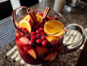Close-up of a glass pitcher filled with vibrant red holiday sangria, loaded with orange slices, cranberries, apple chunks, and cinnamon sticks.