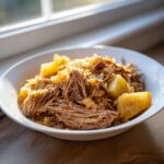 Close-up of shredded pork and sauerkraut mixed with chunks of potato served in a white bowl.