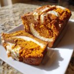 A loaf of moist pumpkin cream cheese bread with one slice cut, showing the rich orange crumb and cream cheese swirl.