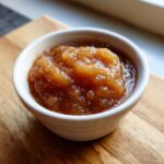 Close-up of rich, brown homemade applesauce served in a small white ramekin on a wooden board.