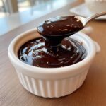 A spoonful of rich, glossy chocolate fondue being lifted from a small white ramekin.