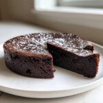 A decadent, dark flourless chocolate cake dusted with powdered sugar, with one slice cut and slightly pulled away.