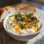 A white bowl filled with creamy ricotta dip, drizzled with chili oil and topped with fresh basil, served with slices of crusty bread.