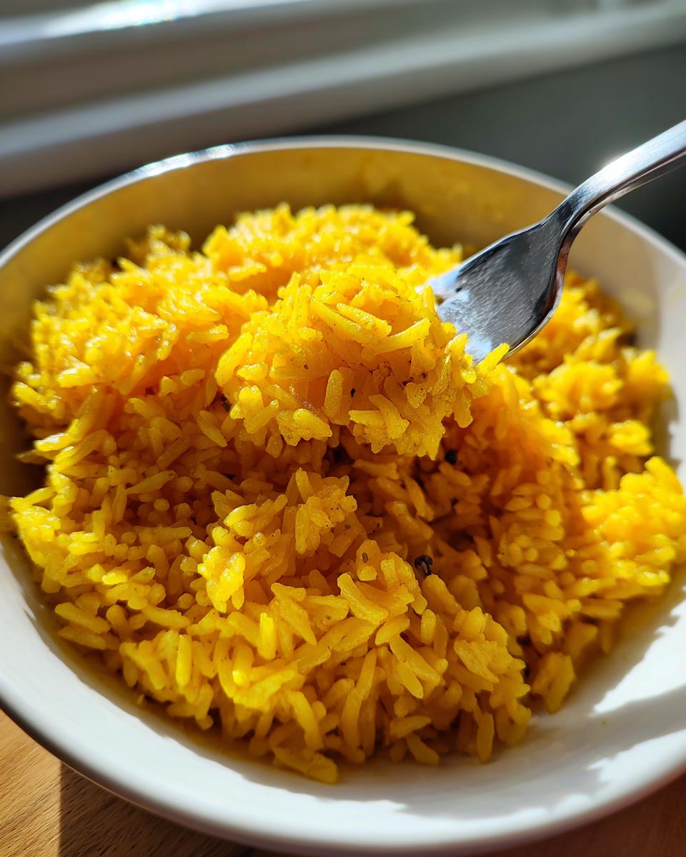 Close-up of fluffy, bright yellow turmeric rice being lifted from a white bowl with a silver fork.