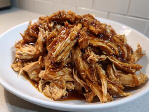 Close-up of shredded crockpot teriyaki chicken piled high on a white plate, glistening with dark teriyaki sauce.