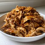 Close-up of shredded crockpot teriyaki chicken coated in glossy sauce, served in a white bowl.