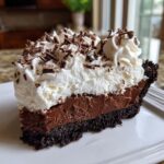 A rich slice of chocolate mud pie featuring a dark crust, thick chocolate filling, and a generous topping of whipped cream and chocolate shavings.