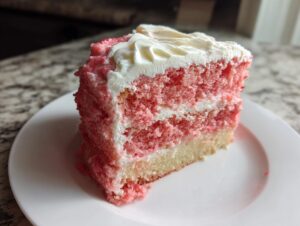Close-up of a moist slice of pink strawberry cake layered with white frosting on a white plate.