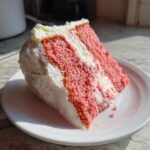 A close-up of a moist slice of pink strawberry cake layered with white frosting on a white plate.