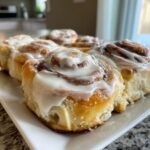 A close-up shot of several freshly baked, soft cinnamon buns generously topped with white cream cheese icing.