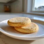 Two round, golden soft sugar cookies stacked slightly on a white plate, topped with coarse sugar crystals.