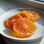 Close-up of four bright orange candied orange slices generously coated in sparkling sugar crystals on a white plate.