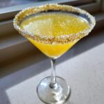 A bright yellow lemon drop martini in a coupe glass with a dramatic gold and silver sugar rim.