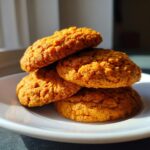 A stack of four golden-orange persimmon cookies resting on a white plate, highlighted by bright sunlight.