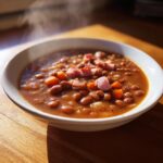 A close-up of a steaming white bowl filled with hearty bean soup, topped with chunks of ham and diced carrots.