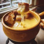 A piece of toasted bread is dipped into a pot of creamy cheese fondue recipe, showing long, irresistible cheese pulls.