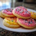 A stack of soft sugar cookies topped with bright pink and yellow sugar cookie icing that hardens, covered in colorful sprinkles.
