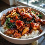 A close-up, appetizing photo of a teriyaki chicken rice bowl featuring glossy, saucy chicken pieces over white rice and green vegetables.