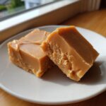 Two golden-brown squares of creamy penuche fudge resting on a white plate near a window.