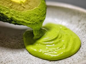 Close-up of a vibrant green avocado sauce being poured from a halved avocado onto a speckled ceramic plate.