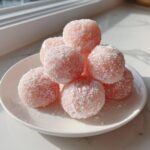 A stack of light pink, round watermelon mochi balls generously dusted with white shredded coconut on a white plate.