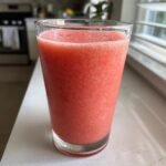 A tall glass filled with a vibrant pink, icy watermelon smoothie sitting on a white kitchen counter.
