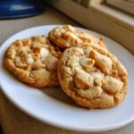 Close-up of three golden brown white chocolate macadamia nut cookies stacked slightly on a white plate.