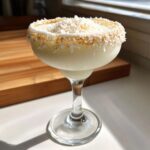 A creamy white Christmas margarita in a coupe glass, garnished heavily with toasted coconut flakes on the rim.