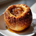 Close-up of a perfectly baked apple coated in cinnamon sugar, sitting in its own syrup on a white plate.