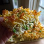 A hand holding up a thick slice of creamy tuna noodle casserole with peas and a crispy topping.