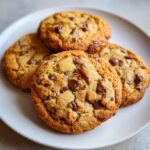 Close-up of four golden brown banana bread cookies loaded with melted chocolate chips on a white plate.