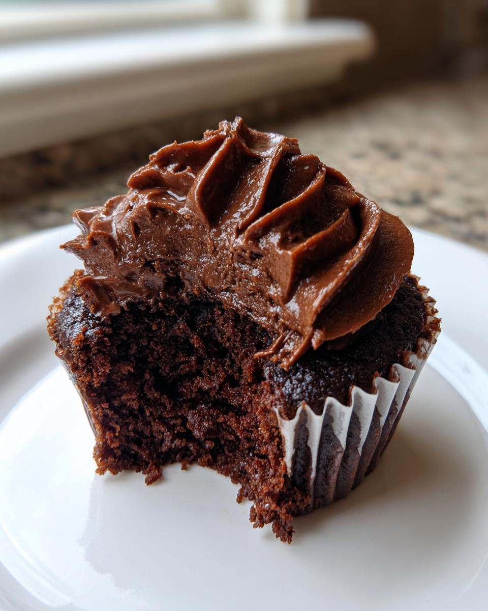 Close-up of a rich, dark chocolate cupcake with a bite taken out, showing moist interior and swirled frosting.