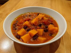 A close-up view of a white bowl filled with rich, orange butternut squash chili containing large chunks of squash and black beans.