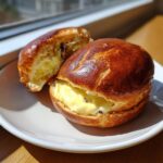 Two halves of a chocolate chip vanilla custard brioche, generously filled with creamy yellow custard, resting on a white plate.