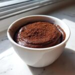 Close-up of rich, dark chocolate protein pudding in a white bowl, dusted heavily with cocoa powder.