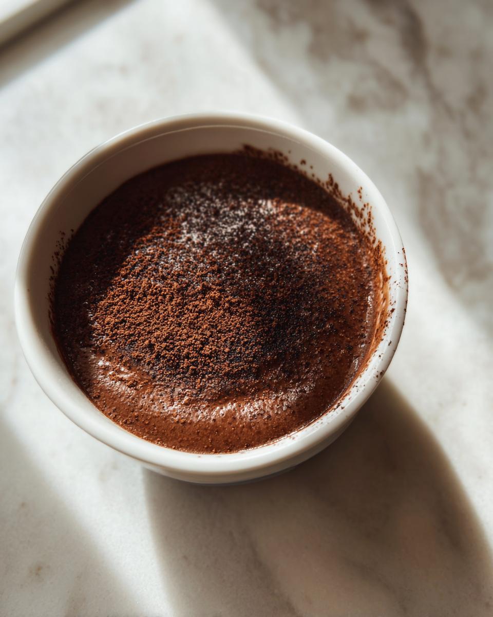 Overhead shot of creamy chocolate protein pudding topped with cocoa powder in a white bowl.