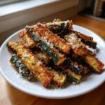 A pile of golden brown, crispy air fryer zucchini sticks topped with grated Parmesan cheese on a white plate.