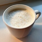 Close-up of a mug filled with creamy, foamy white hot chocolate, catching warm sunlight.
