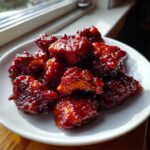 A white plate piled high with pieces of glossy, dark red gochujang chicken, catching the light near a window.