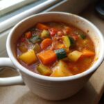Close-up of a white mug filled with chunky, tomato-based vegetable soup featuring carrots, zucchini, and potatoes.