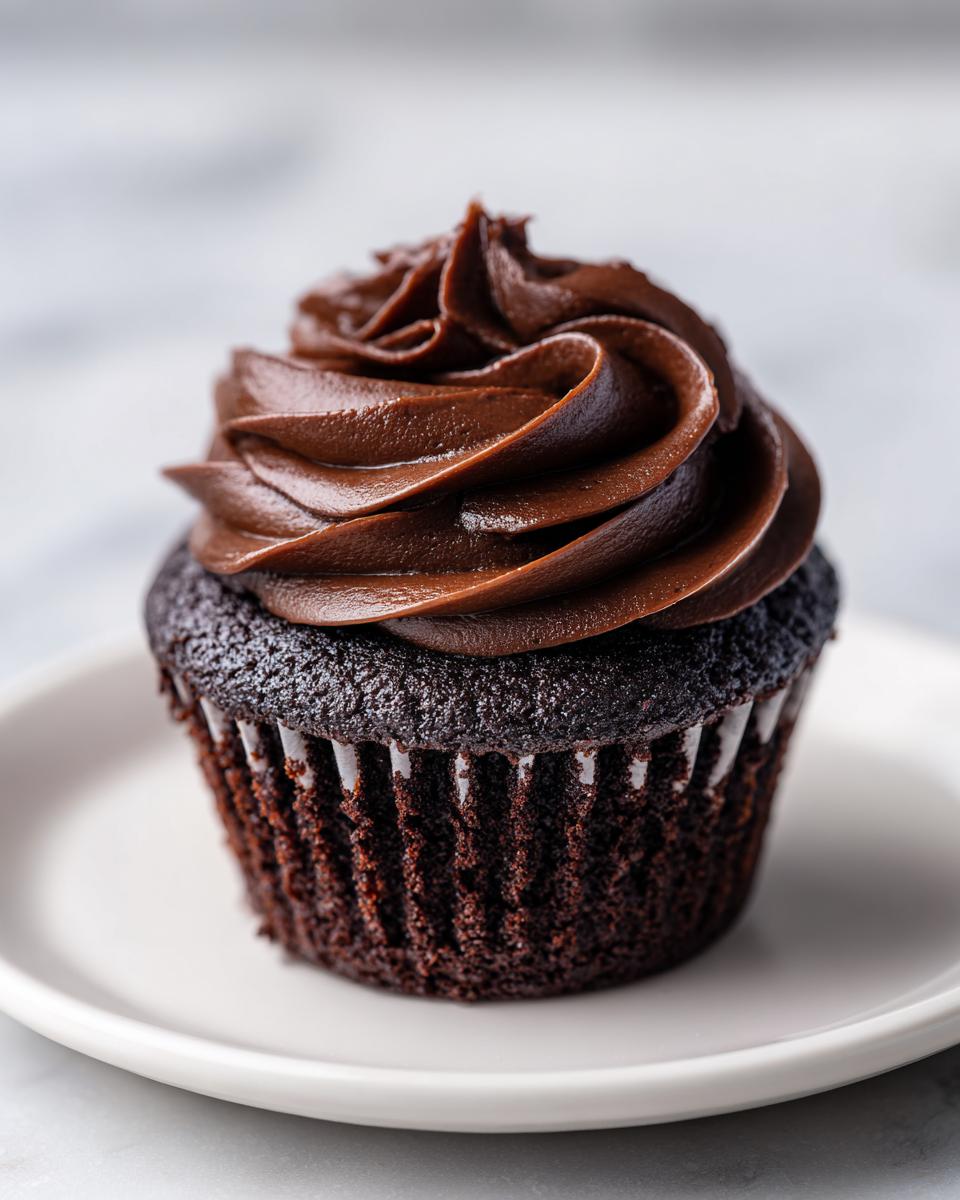 A single, rich, dark chocolate cupcake topped with a generous swirl of glossy chocolate frosting, sitting on a white plate.