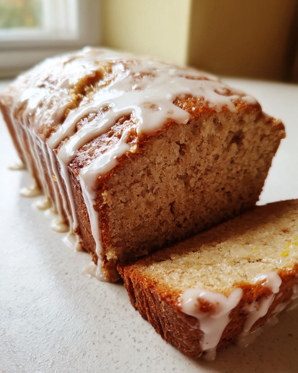 A close-up of moist eggnog bread loaf with a thick vanilla glaze dripping down the sides, one slice cut.