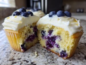 Close-up of a lemon blueberry cupcake cut in half showing moist yellow cake filled with burst blueberries and topped with cream cheese frosting.