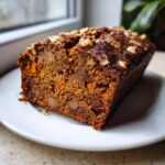 A close-up of a moist slice of persimmon bread topped with chopped walnuts, sitting on a white plate.