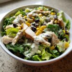 A bowl filled with a fresh southwest salad featuring lettuce, grilled chicken strips, black beans, corn, and creamy dressing.